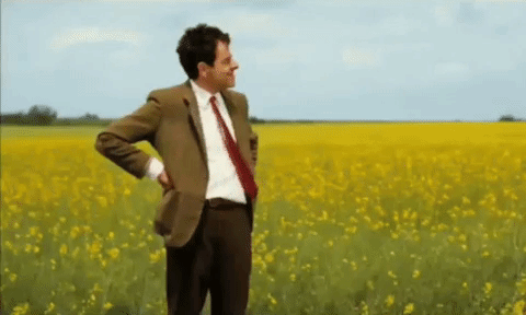 Mr. Bean waiting ni front of a yellow field of flowers, checking his watch'